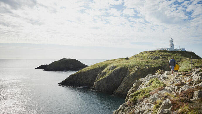 Man and child walking on a coastal path towards a lighthouse