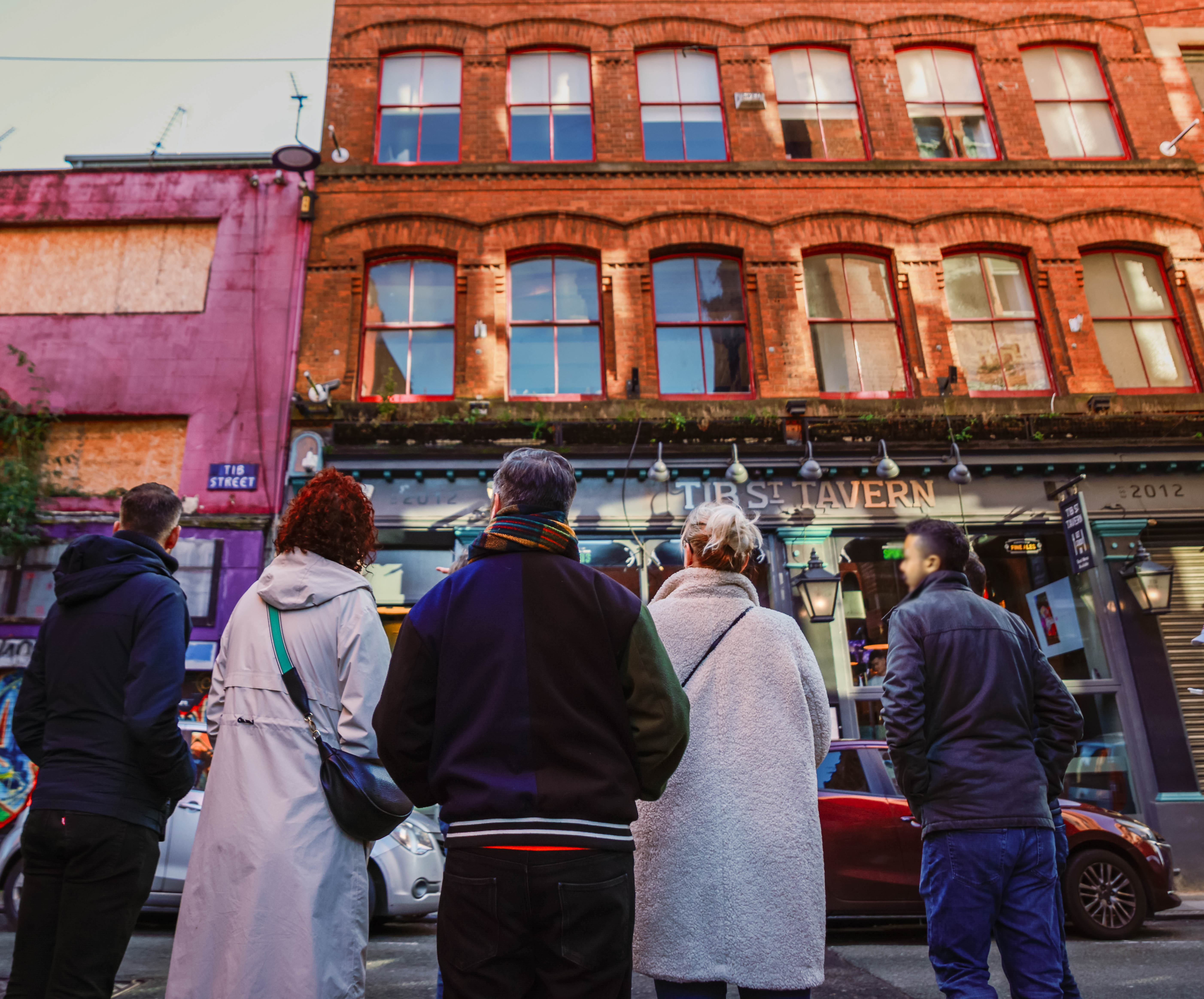 Five people stand on a street facing a red-brick pub called Tib Street Tavern in an urban area with colorful buildings.