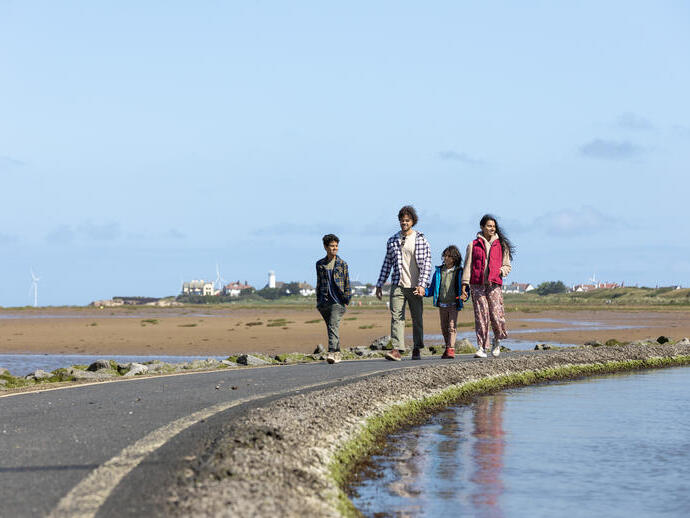 A group of people walking along a coastal path with sandy beach, water, wind turbines, and buildings in the background.