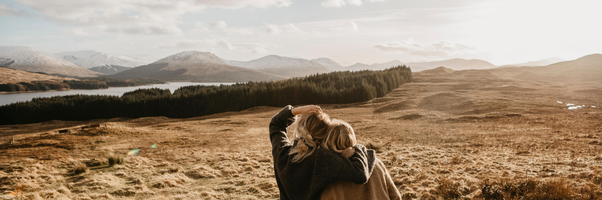 Zwei Freunde umarmen sich und blicken über das Tal auf einen See und die Berge