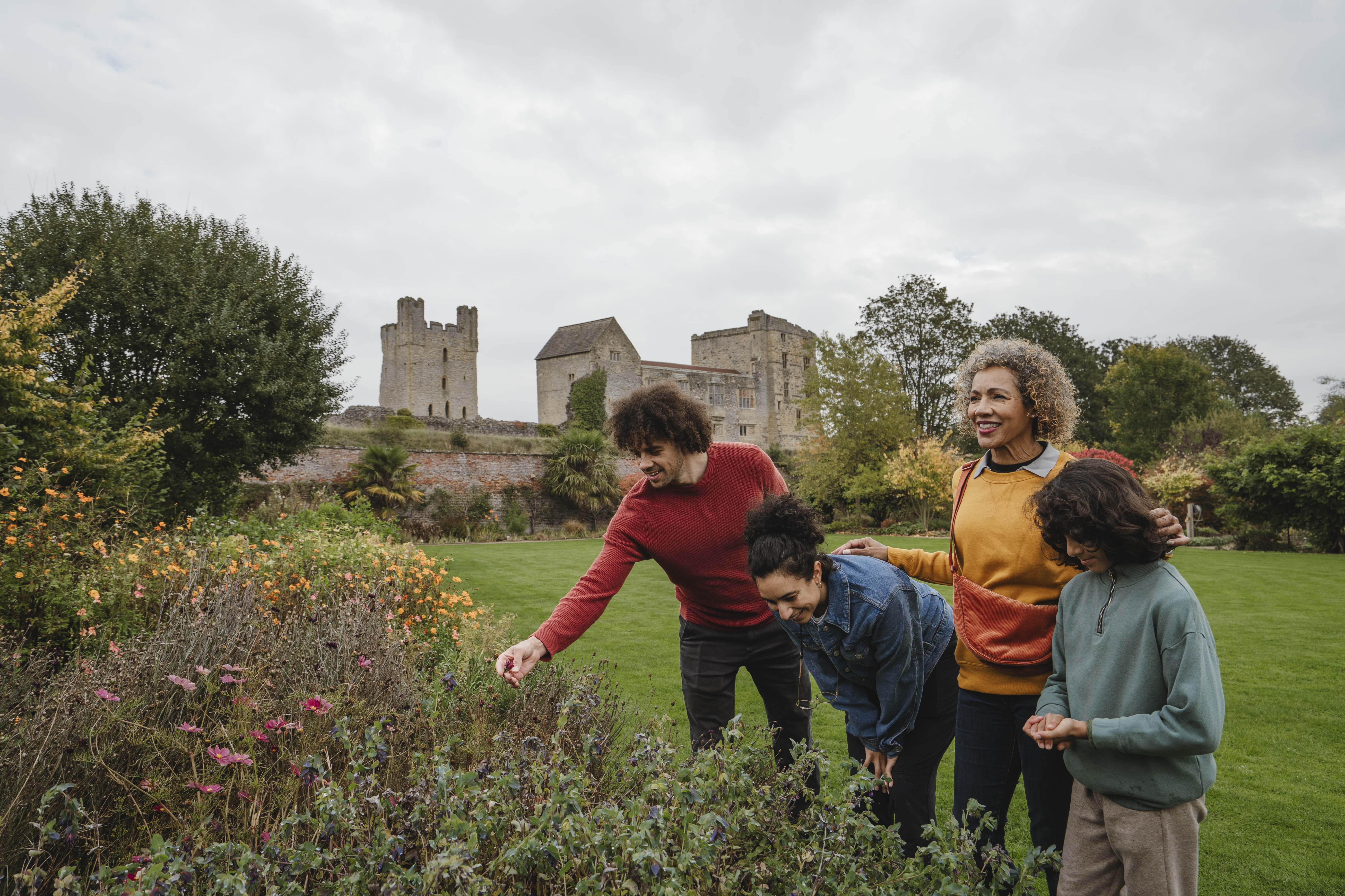 A group enjoys a flower garden with a historic stone castle and green lawns in the background on a cloudy day.