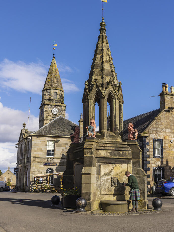 A man gets water from a large stone fountain with faded red lion statues in a town square.