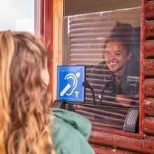 A person inside a ticket booth smiles at someone standing outside. A blue hearing loop sign is prominently displayed. Noahs Ark Zoo Farm - Gold award winner for the Accessible and Inclusive Tourism Award at the VisitEngland Awards for Excellence 2023.