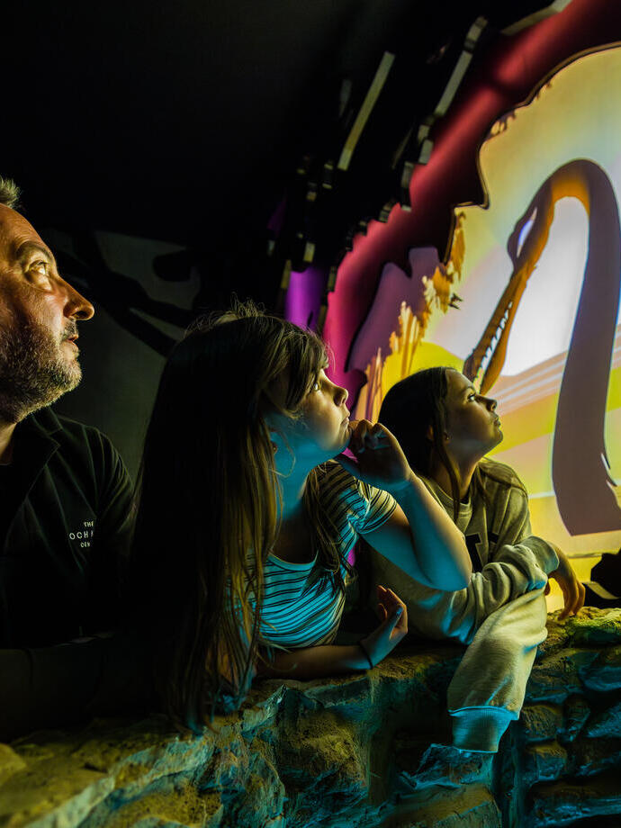 A man and two young girls watch an animated water monster with a long neck on a screen .