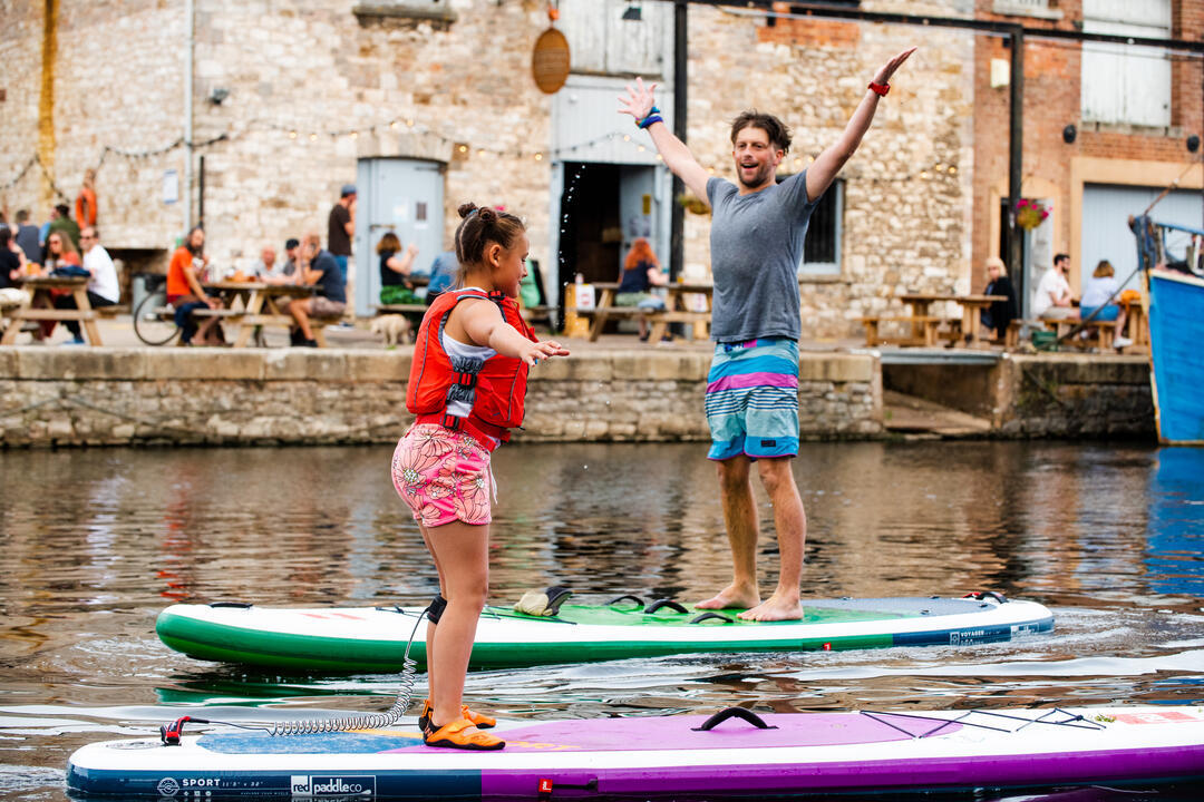 Ein Kind und ein Mann stehen auf Paddleboards in einem Fluss in einer städtischen Umgebung