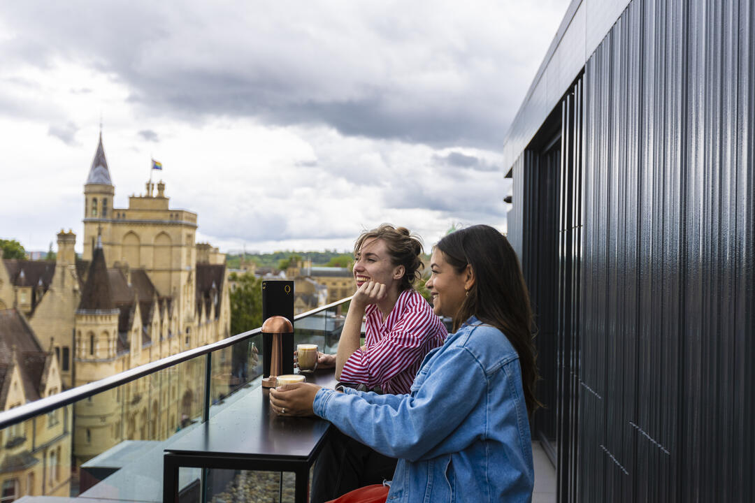 Two women drink coffee on a rooftop overlooking the city