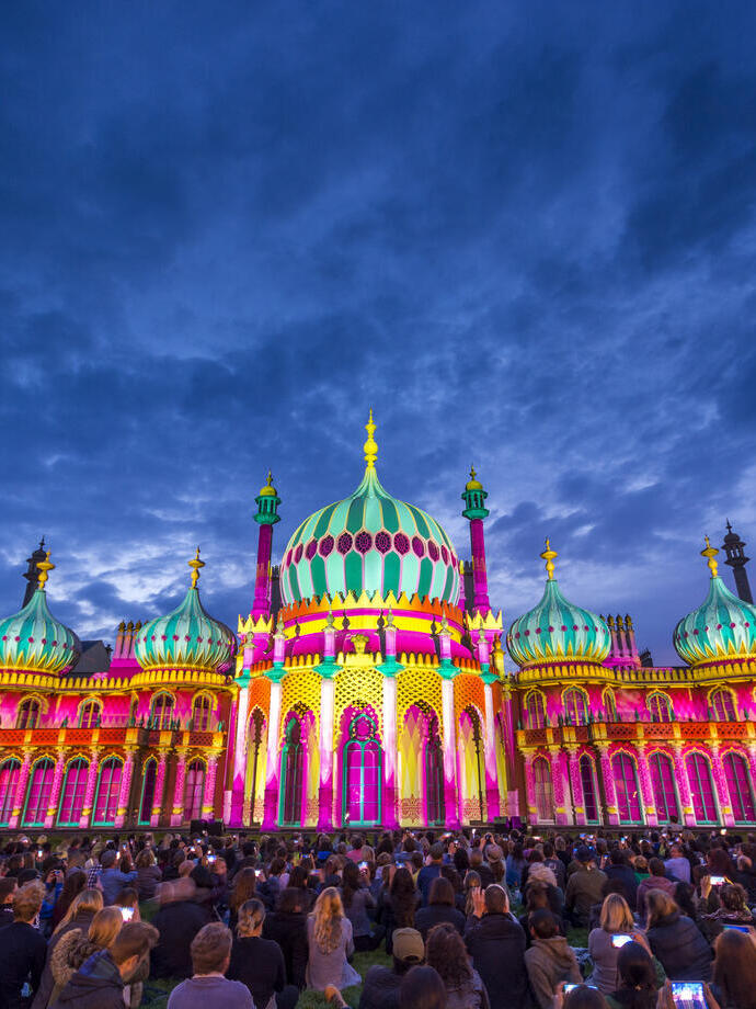 Crowd sitting on the ground in front of a large ornate building lit up with bright colours