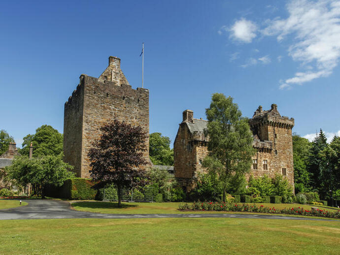 Außenansicht eines kleinen Schlosses mit Fahnenmast und einigen Bäumen an einem sonnigen Tag.