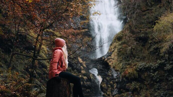 Woman in a red hat and jacket sits on a tree stump looking up at the Pistyll Rhaeadr waterfall