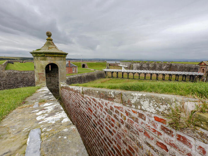 A brick defensive wall with a small tower amid a larger stone fortification under grey skies.