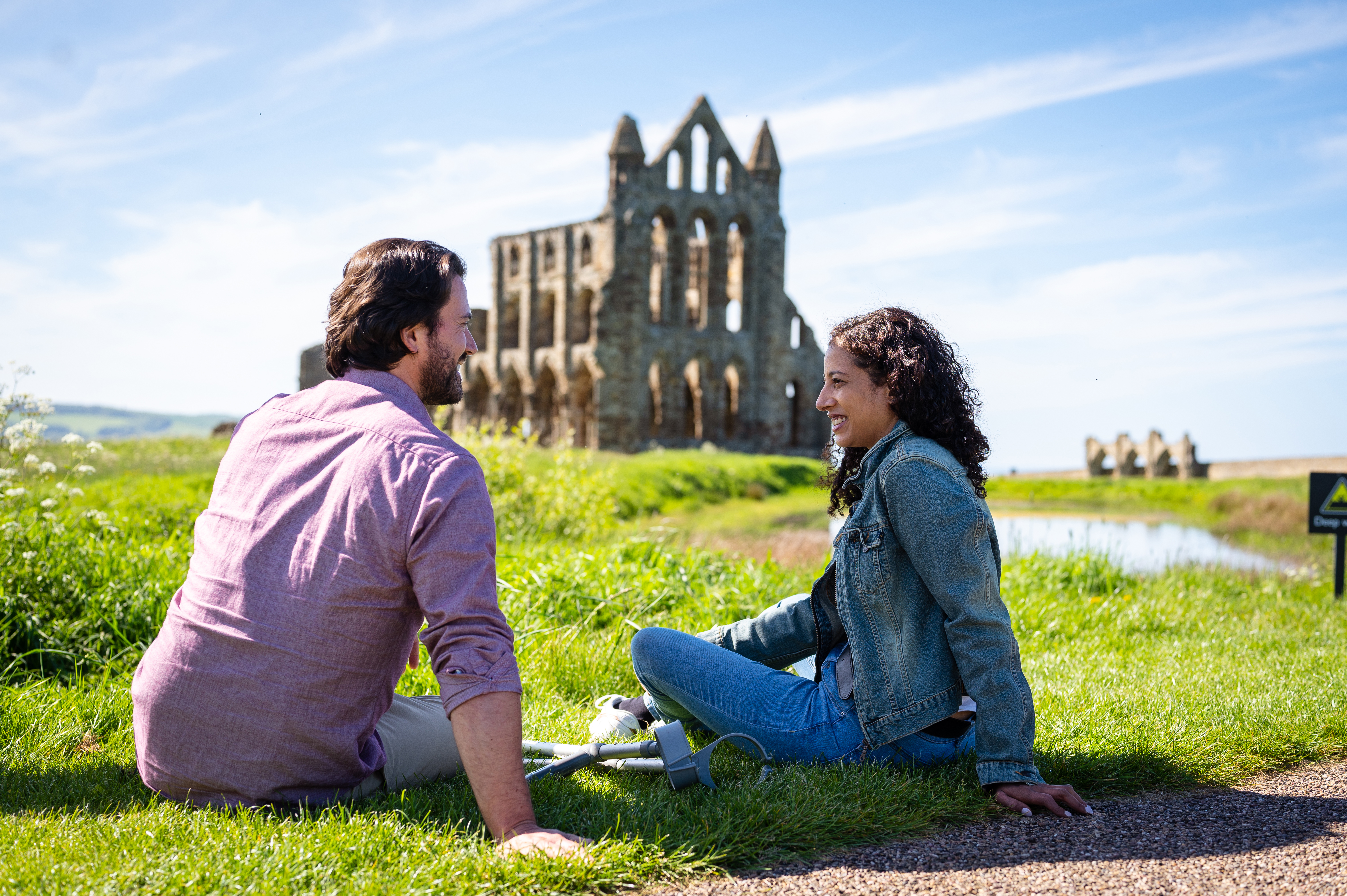 Man and woman sitting on grass with crutches beside them, an Abbey in the background
