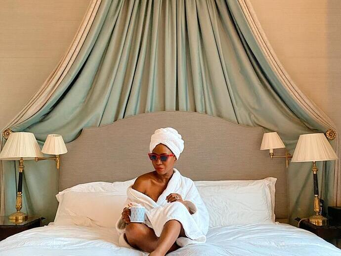 Une femme assise sur un lit, vêtue d'un peignoir et portant des lunettes de soleil, dans une chambre d'hôtel