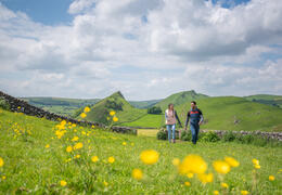 Two people walking through a green meadow with yellow flowers, hills, and stone walls under a partly cloudy sky.