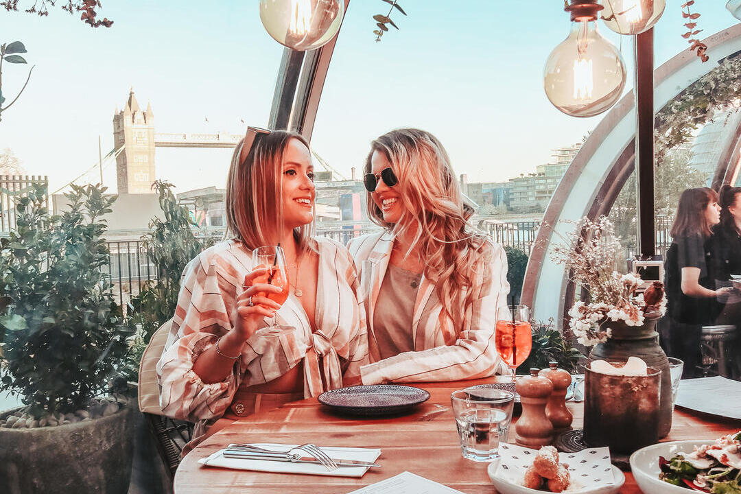 Two women enjoying food and drink in a glass dining pod on the river side