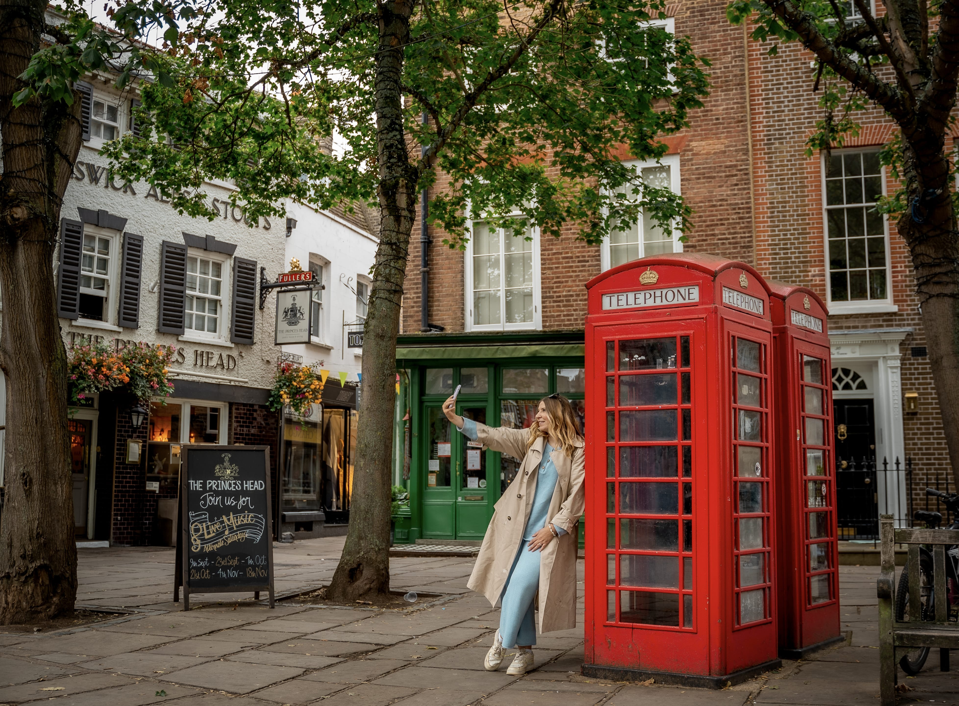 A woman leaning against a red telephone box and takes a selfie
