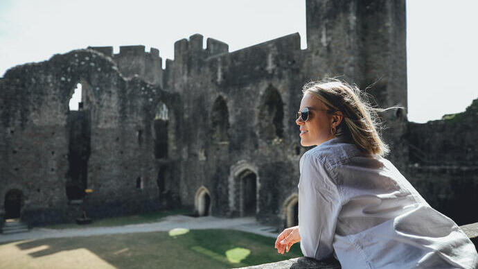 Persona con gafas de sol apoyada en una repisa de piedra en las ruinas de un castillo histórico, con luz solar y ventanas arqueadas al fondo.