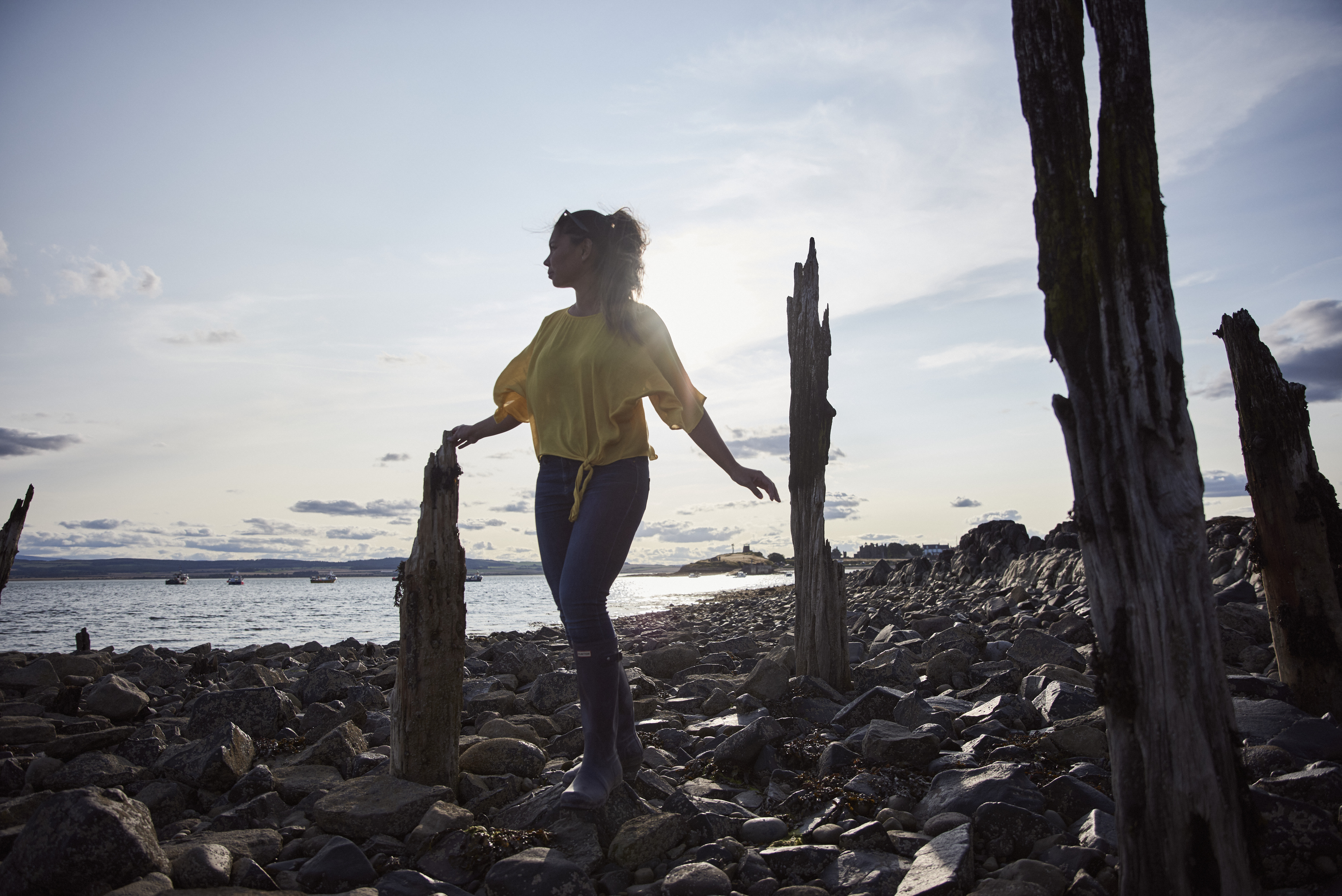 Woman walking on large stones in between old wooden stakes by the sea