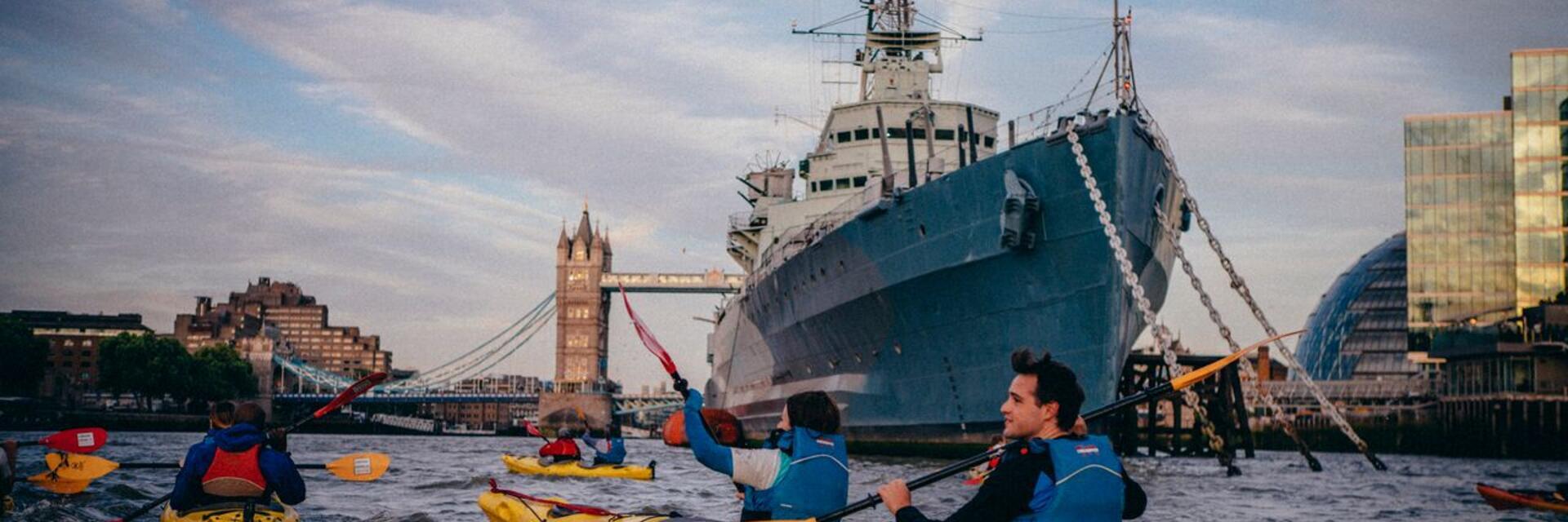 Personas remando en kayaks amarillos por un río, frente a un gran barco y un puente, con edificios al fondo.