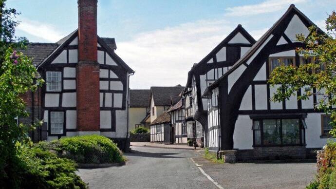 Street with traditional black and white Tudor-style buildings under a blue sky, surrounded by greenery and quiet atmosphere.