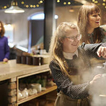 A young woman working the coffee machine with colleague assisting in cafe