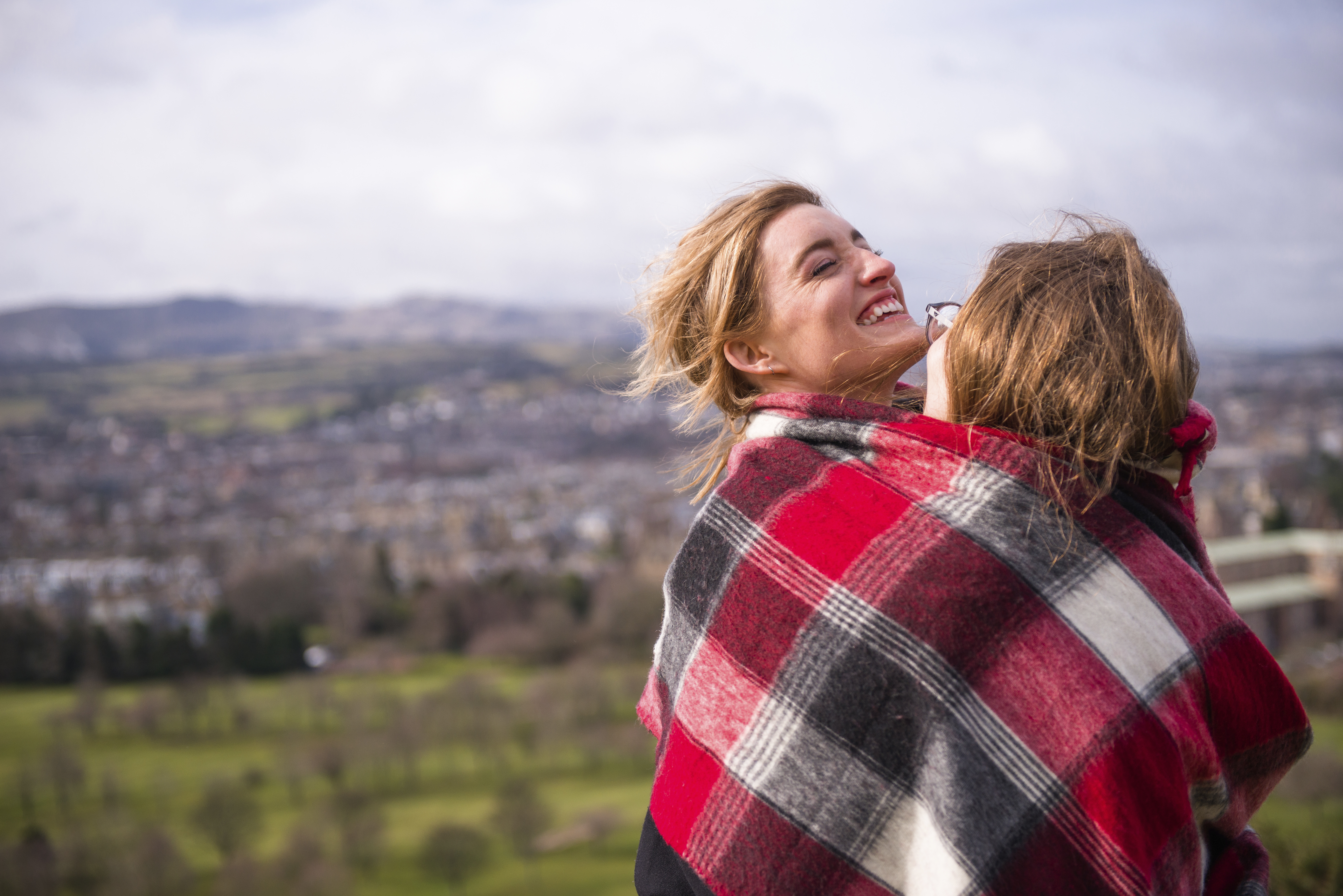 Two women wrapped in red checkered blanket standing on a hill overlooking a city