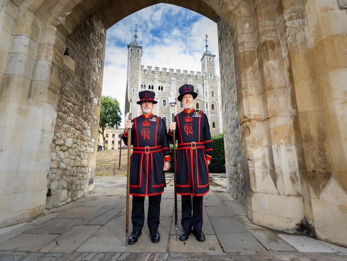 Two people in traditional Beefeater uniforms stand at a stone gate with the Tower of London in the background under a blue sky.