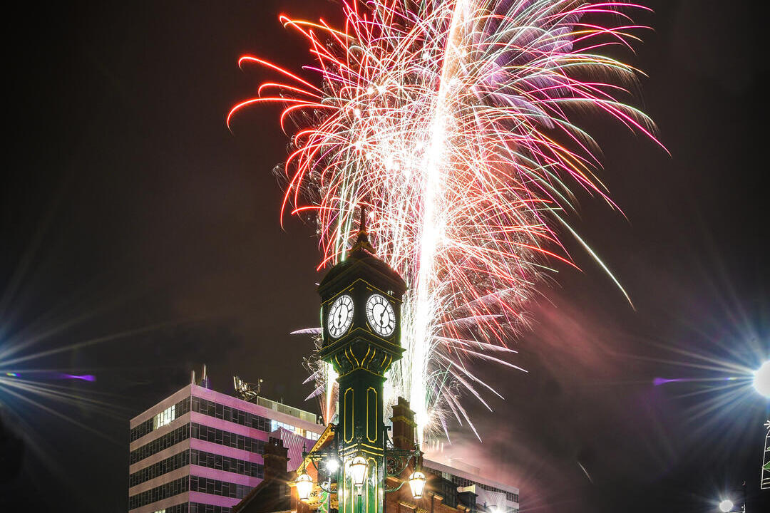 A historic green clock tower stands in a city center at night with vibrant fireworks lighting the sky behind it.