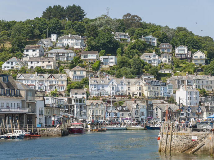 Seaside village with hillside houses, lush greenery, a harbour with boats, and waterfront buildings under a clear blue sky.