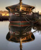 Historic wooden ship docked at a harbor, reflected in calm water with a ramp to the side and buildings in the background at sunset.