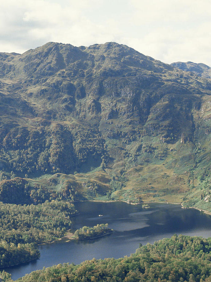 Vue lointaine d'un loch entouré d'arbres et de montagnes verdoyantes.