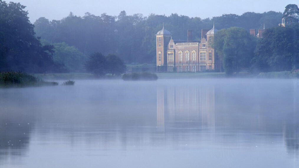 Früher Morgen auf dem Anwesen Blickling Hall, mit dem Haus, das sich im Wasser des Sees spiegelt, und einem bläulichen Nebel ringsum