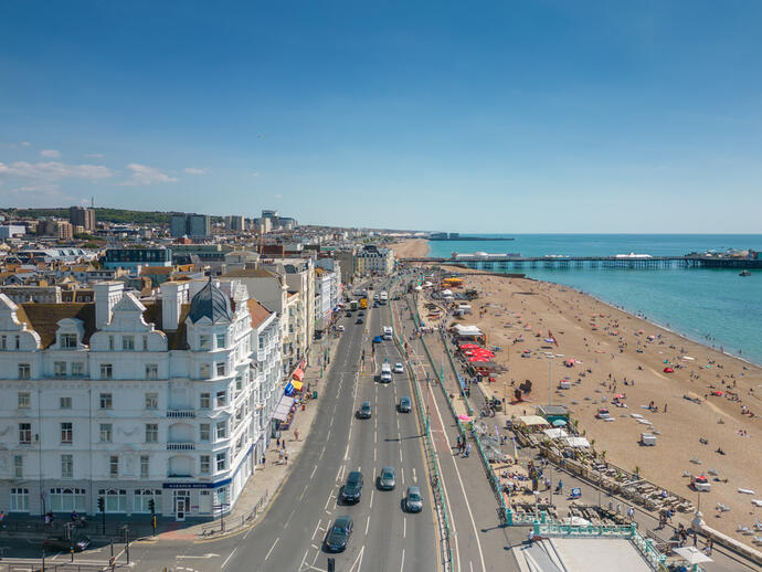 A view of a busy seaside city with a sandy beach, pier, seafront road, historic buildings, and people enjoying a sunny day.