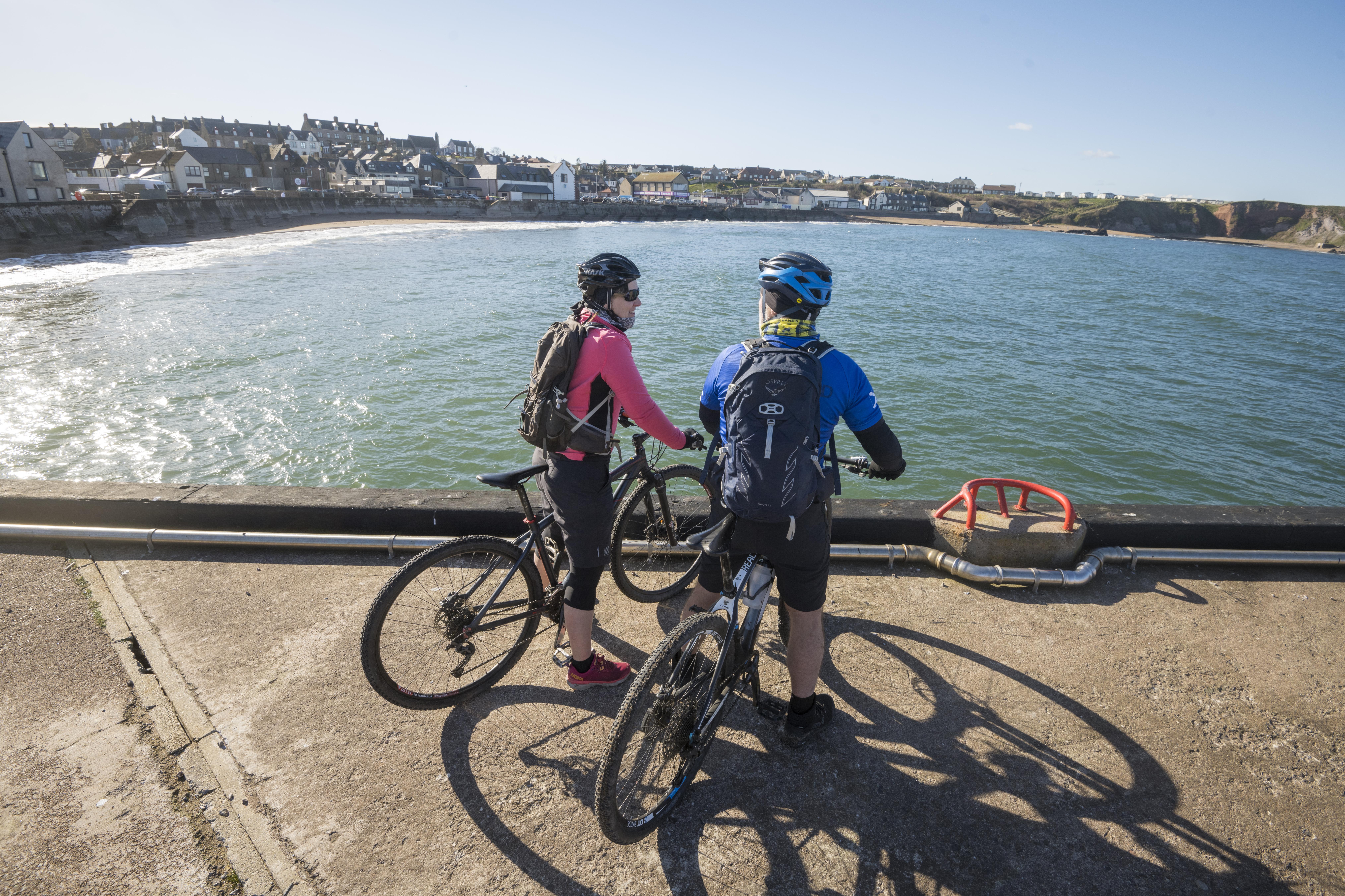 Two cyclists pause by the seaside, looking toward a coastal town with houses and cliffs under a clear sky.