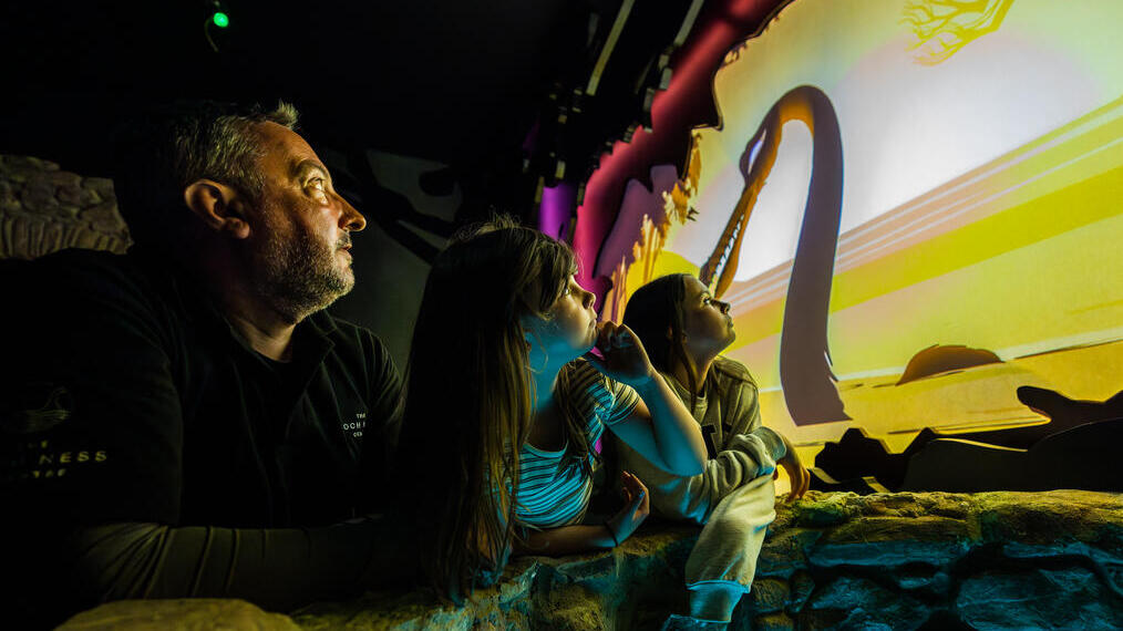 A man and two young girls watch an animated water monster with a long neck on a screen .