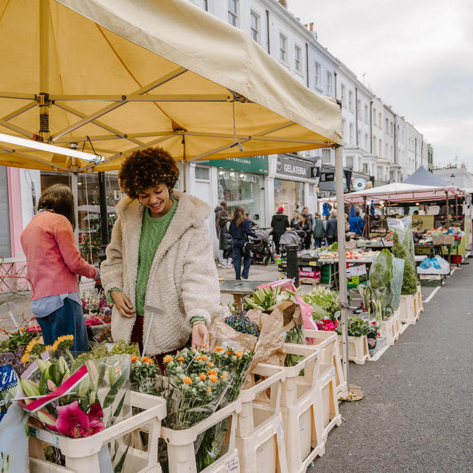 Outdoor flower market scene on a city street with people shopping at stalls under yellow tents, surrounded by historic buildings.