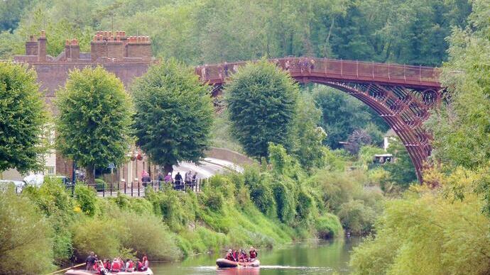 People rafting on a river below a historic iron bridge, with greenery, trees, and brick buildings in the background.