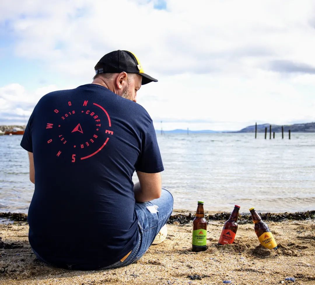 A man sat on a beach next to bottles of Snowdon Craft Beer
