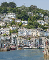 Seaside village with hillside houses, lush greenery, a harbour with boats, and waterfront buildings under a clear blue sky.