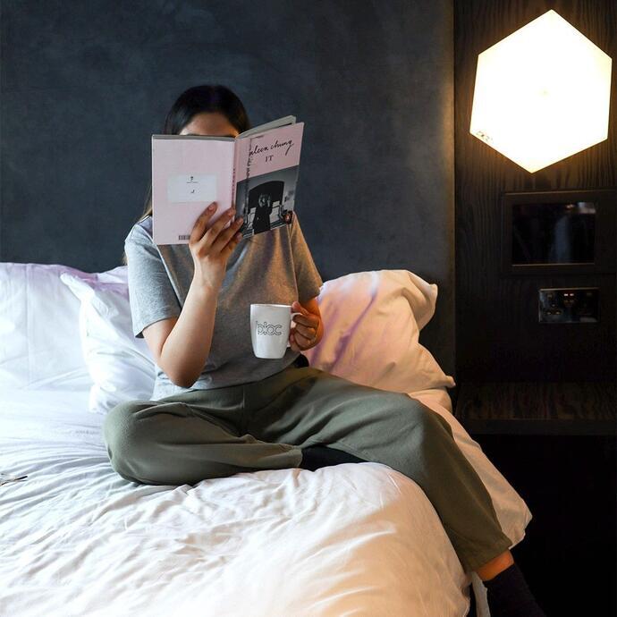 Mujer sentada en la cama de un hotel leyendo un libro y tomando una bebida caliente
