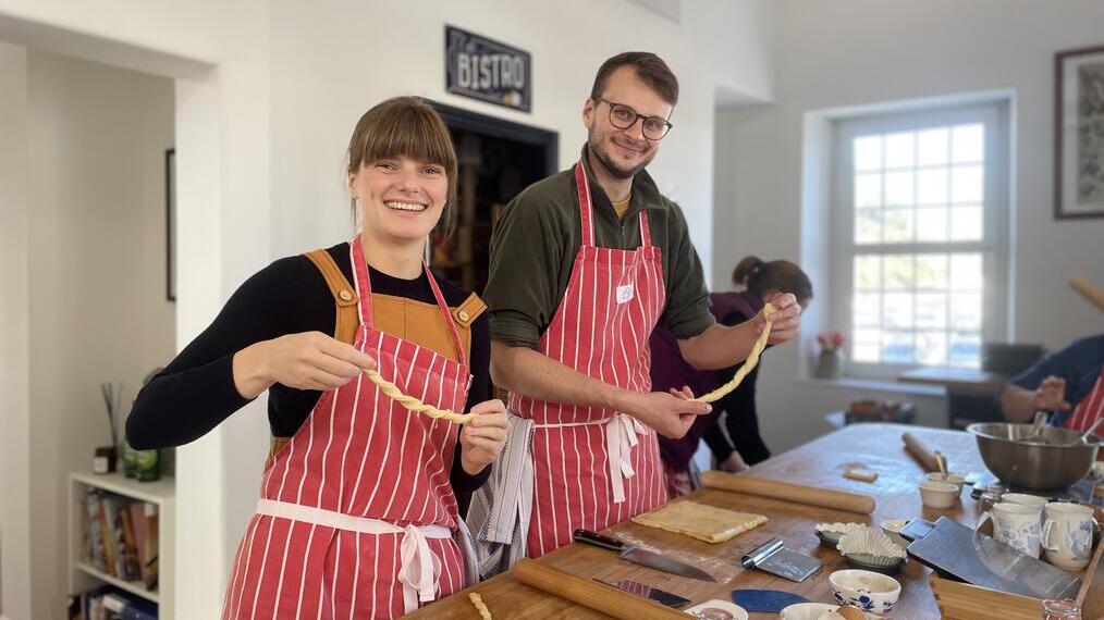 Two people in striped aprons smiling and twisting dough in a bright kitchen, with baking tools and ingredients on the table.