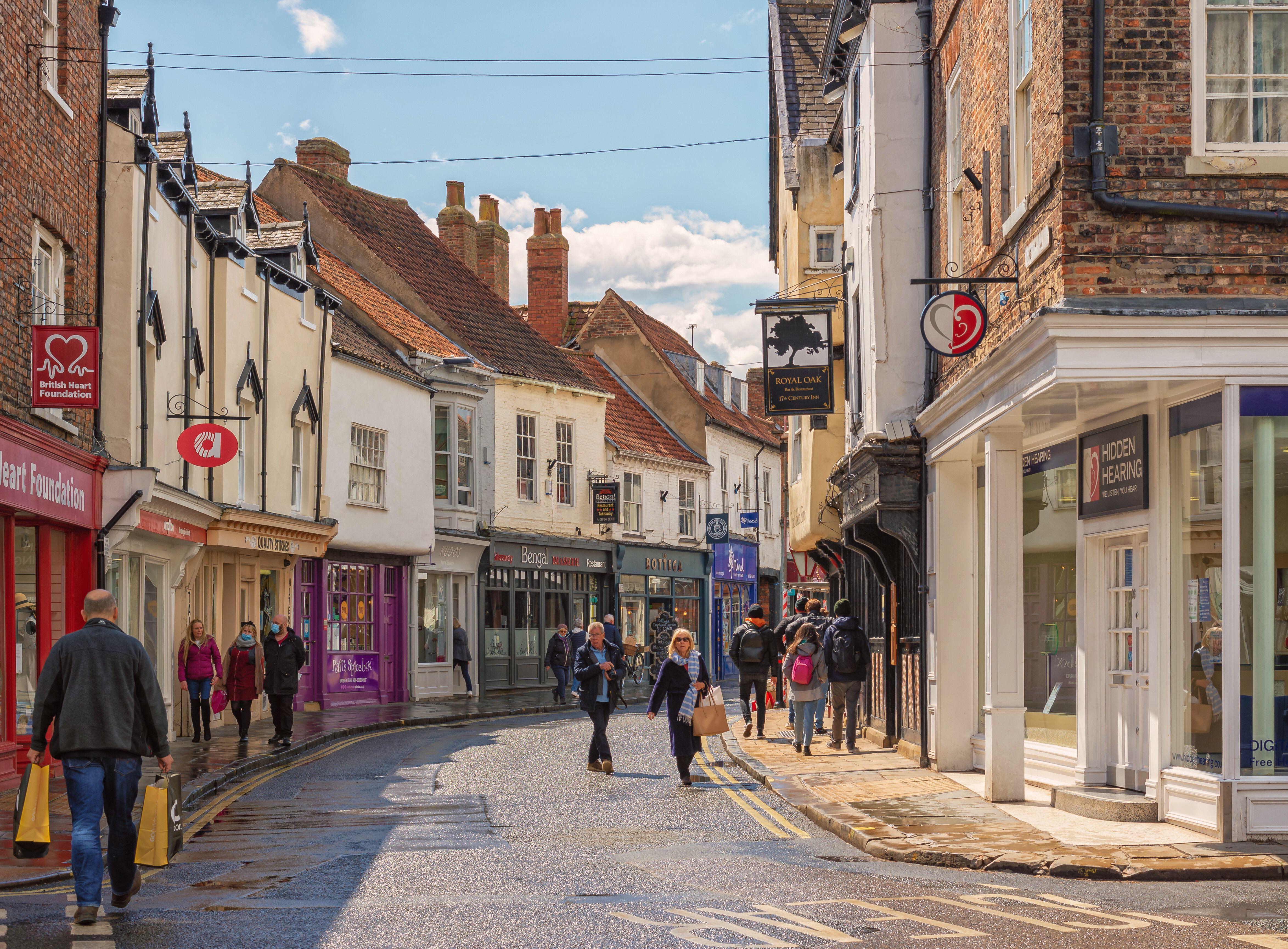 Shopping street just after a rain shower. Historic buildings line the street and shoppers walk between them. A sky with cloud is overhead.
