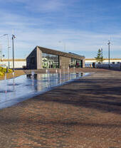 Modern waterside plaza with small fountains, a triangular building, landscaped greenery, and people walking under a clear blue sky.