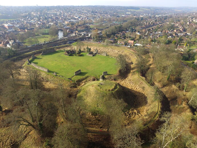 Aerial view of a historic castle ruin with stone walls and a central mound, surrounded by trees and town buildings in the background.