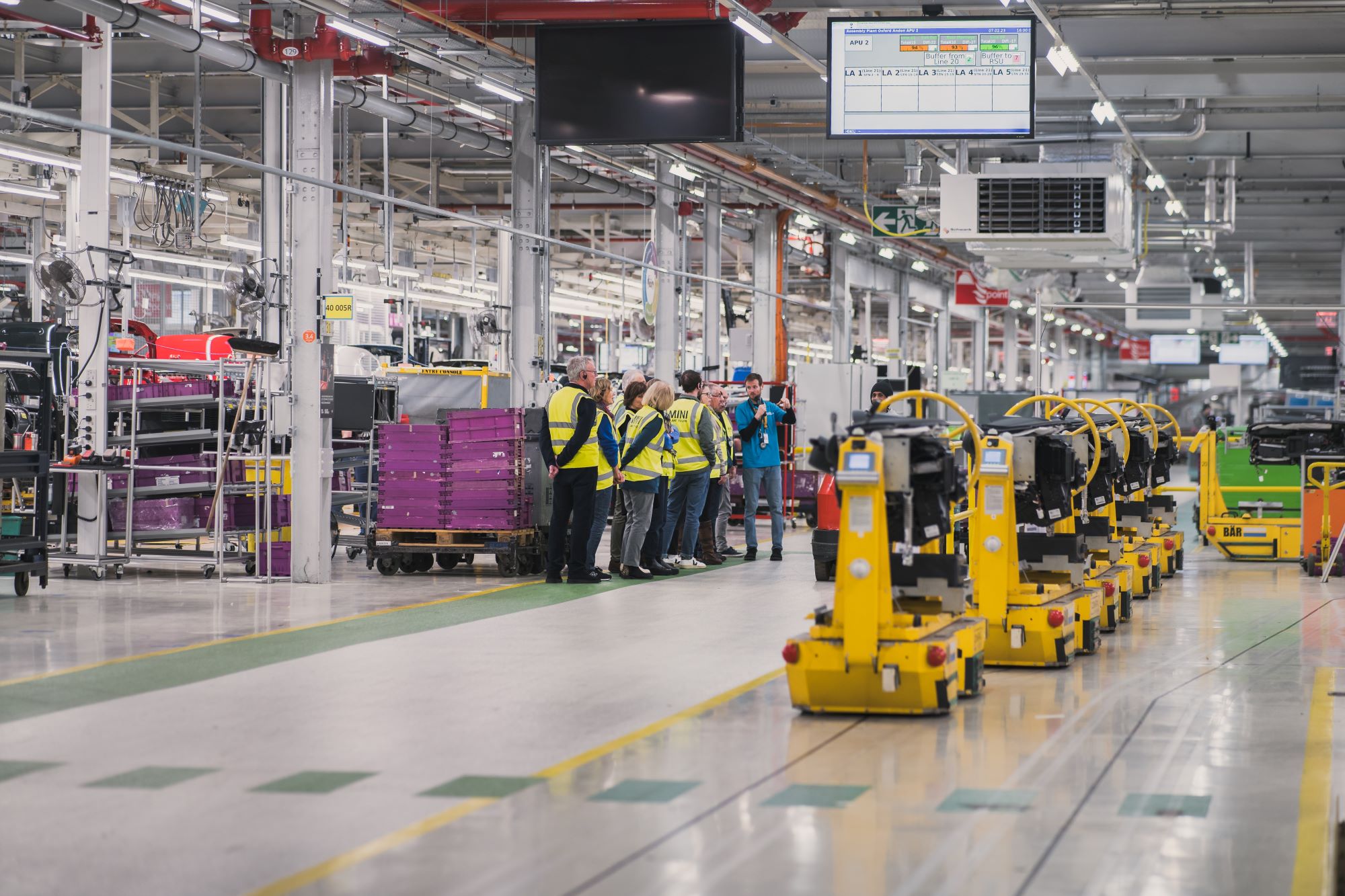 People in safety vests tour a modern factory with machinery, assembly lines, and overhead digital displays.