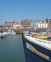 Harbor with sailboats and a large boat in foreground, colorful waterfront buildings under a clear blue sky.