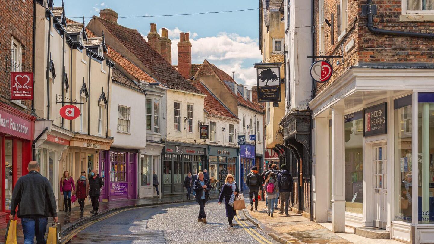 Einkaufsstraße kurz nach einem Regenschauer. Historische Gebäude säumen die Straße, zwischen denen die Einkäufer hindurchgehen. Über ihnen ist ein bewölkter Himmel zu sehen.