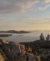 Zwei Männer sitzen auf einer Felsklippe und blicken in der Abenddämmerung auf das Meer