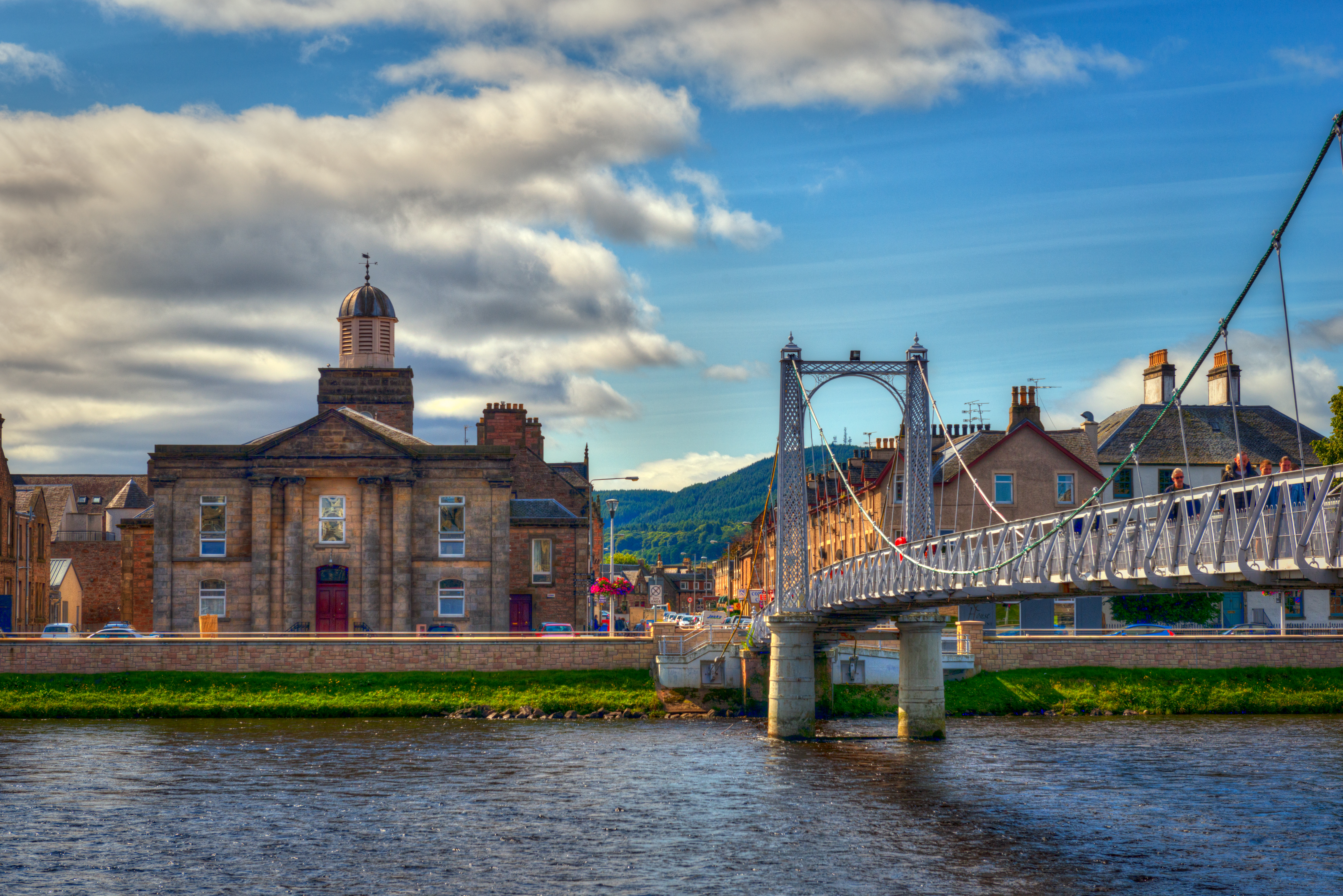 A suspension bridge with people walking across spanning a wide river with houses on the other side