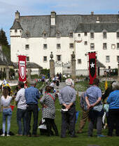 A crowd enjoys an outdoor event in front of a large historic castle, with medieval banners and tents on the grounds.