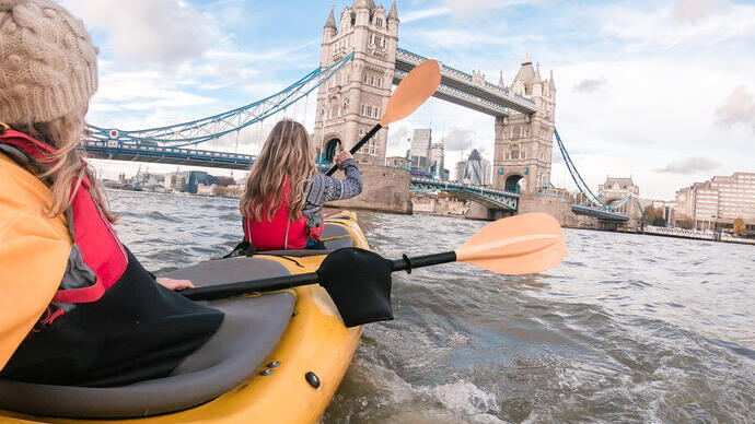 Two women paddling in a kayak on a river towards a bridge in a city